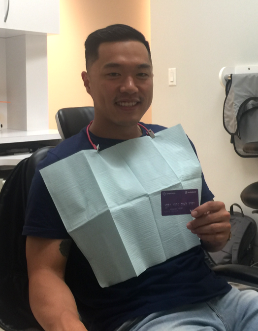 Young man sitting in a dental chair with bib on smiling while holding an Opencare visa giftcard